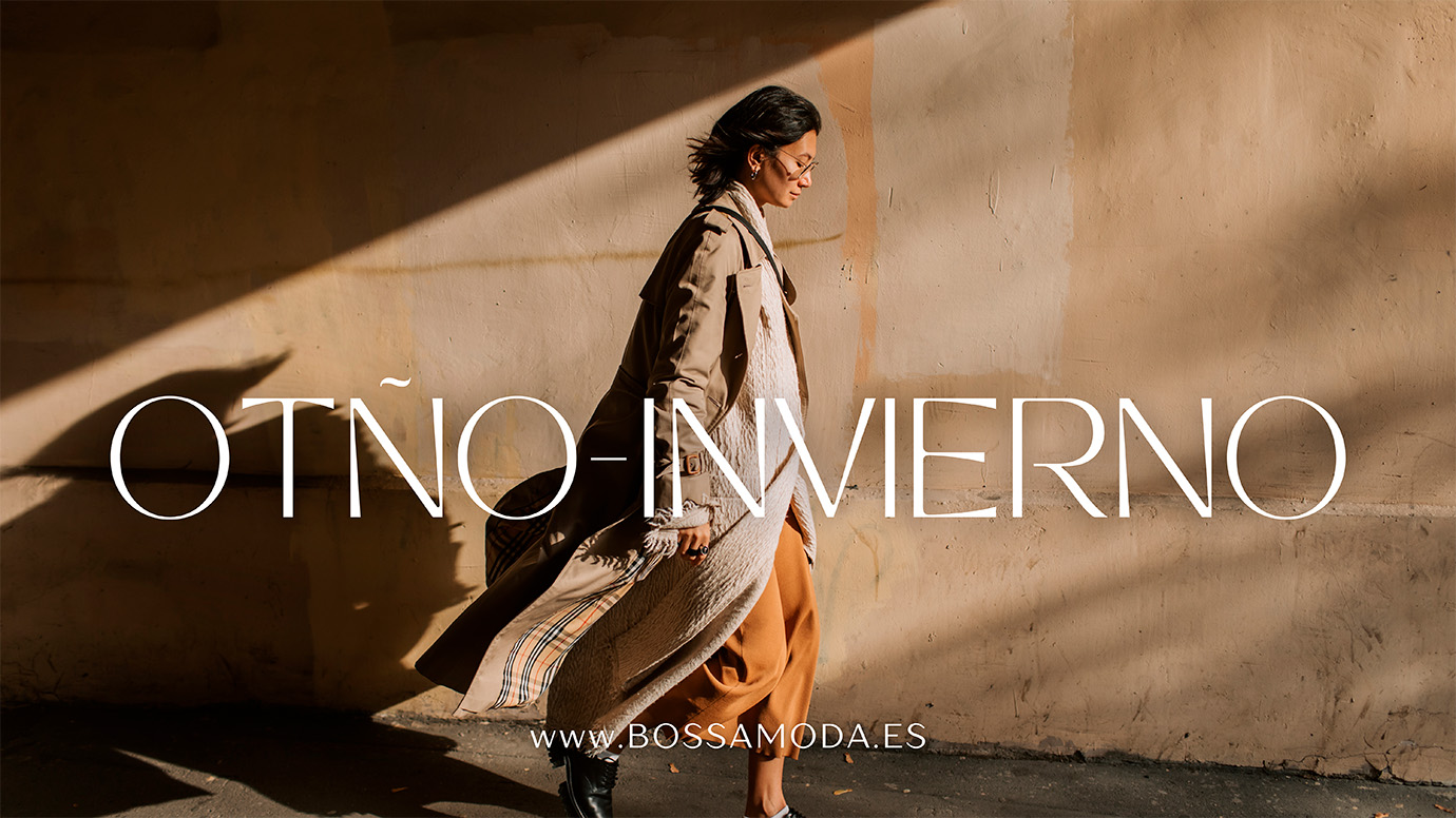 La imagen muestra a una mujer caminando junto a una pared iluminada parcialmente por la luz solar, lo que genera sombras y contrastes cálidos. La mujer lleva un abrigo largo beige y un conjunto de tonos otoñales, complementado con gafas y un bolso discreto. El texto en la imagen dice "OTOÑO-INVIERNO" en letras grandes y elegantes, con la dirección web "www.bossamoda.es" en la parte inferior. La composición evoca un estilo sofisticado y moderno, ideal para destacar tendencias de moda para las temporadas frías.