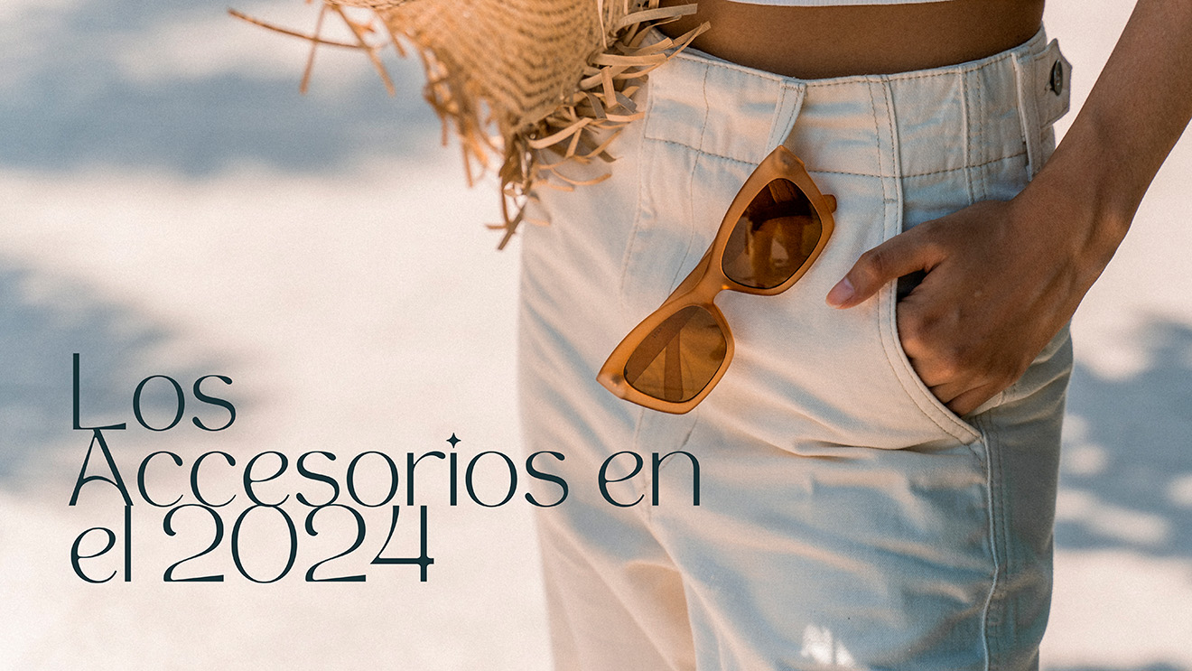 La imagen es una fotografía artística que muestra la portada de un artículo sobre accesorios de moda para el año 2024. En la foto, se ve la cintura de una persona vestida con pantalones claros y un top blanco, que sostiene un sombrero de paja en la mano derecha. Unas gafas de sol de color marrón, que son parte de las tendencias de moda 2024, están colgadas en el bolsillo delantero del pantalón. El fondo desenfocado, con sombras suaves, resalta el enfoque en los accesorios de moda.